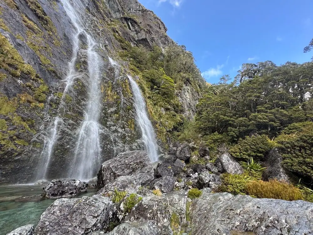 The Tall Earland Falls cascading down a rocky cliff into a clear pool surrounded by mossy boulders and forest on the Routeburn Track. Bright blue sky peeks through above the dramatic landscape.