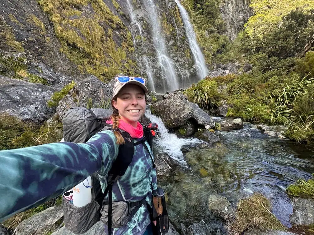 Smiling hiker wearing a backpack takes a selfie beside Earland Falls and a rocky stream along the Routeburn Track. Clear rushing water and mossy cliffs create a dramatic alpine backdrop.