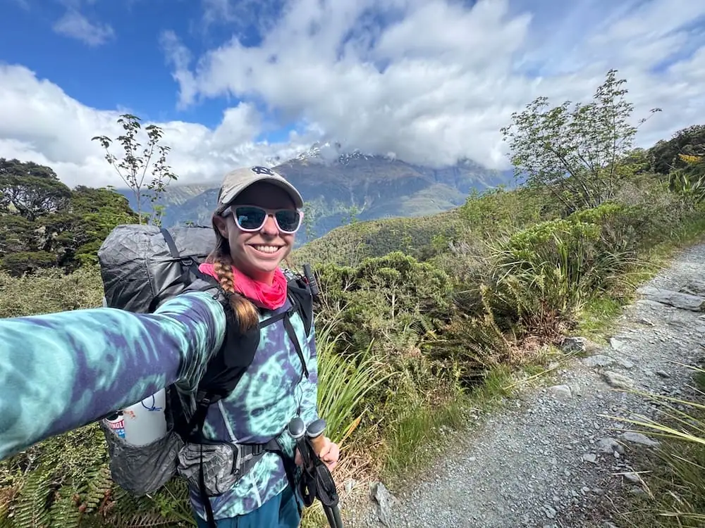 Smiling hiker with trekking poles and a backpack pauses for a selfie beside a gravel trail on the Routeburn Track. Mountains covered in low clouds rise behind dense alpine shrubs.