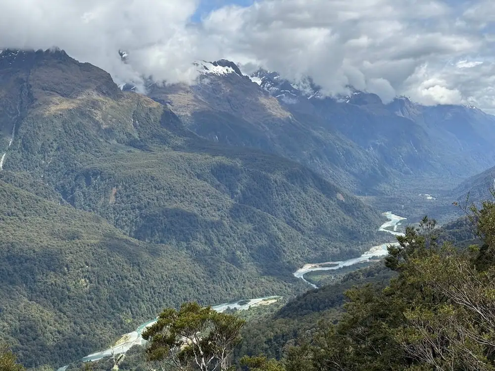 Elevated view of a winding pale blue river flowing through a deep mountain valley on the Routeburn Track. Low clouds drift across steep forested slopes and snow dusted peaks.