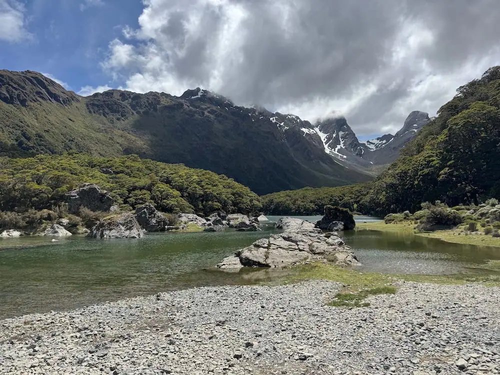 Quiet alpine lake bordered by rocky shoreline and forested mountains along the Routeburn Track. Dark clouds hang above rugged peaks while calm green water reflects the surrounding landscape.