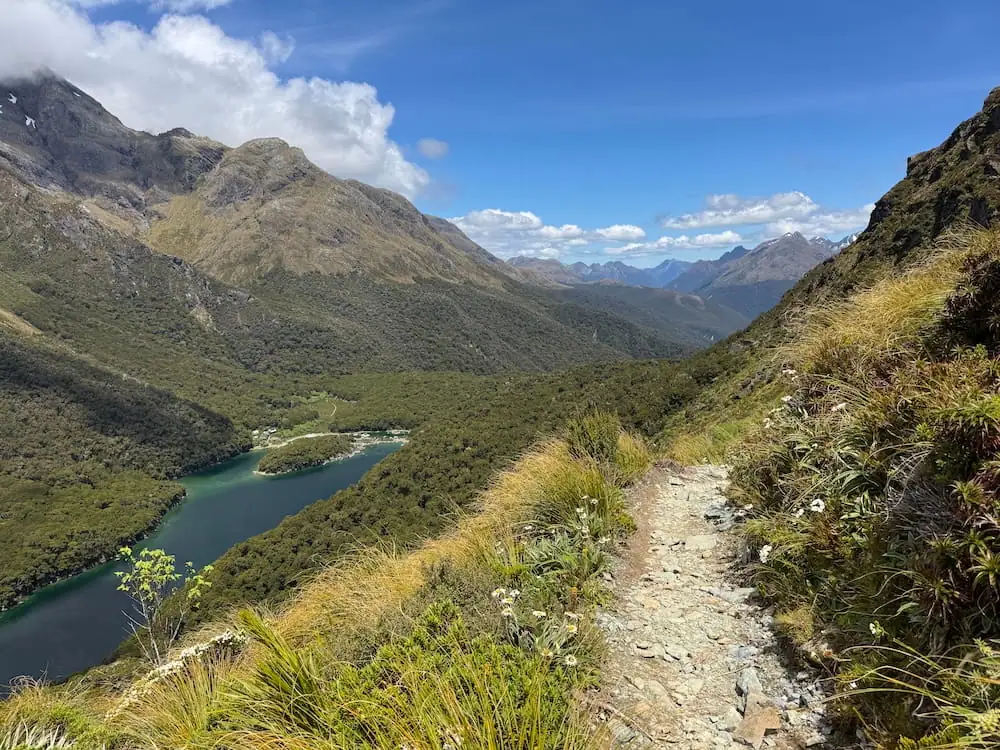 Narrow rocky trail winding along a steep mountainside above a turquoise lake on the Routeburn Track. Rolling mountain ridges stretch into the distance beneath a clear blue sky.