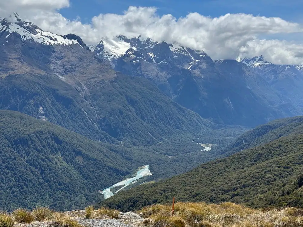 Sweeping mountain valley view with a winding turquoise river cutting through dense forest on the Routeburn Track. Snow capped peaks rise in the distance beneath thick drifting clouds.