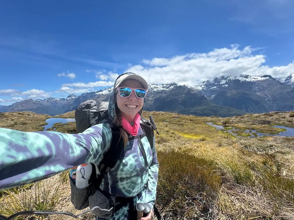 Smiling hiker with a backpack takes a selfie in open alpine terrain on the Routeburn Track. Small reflective ponds and snow capped mountains stretch across the background beneath a bright blue sky.