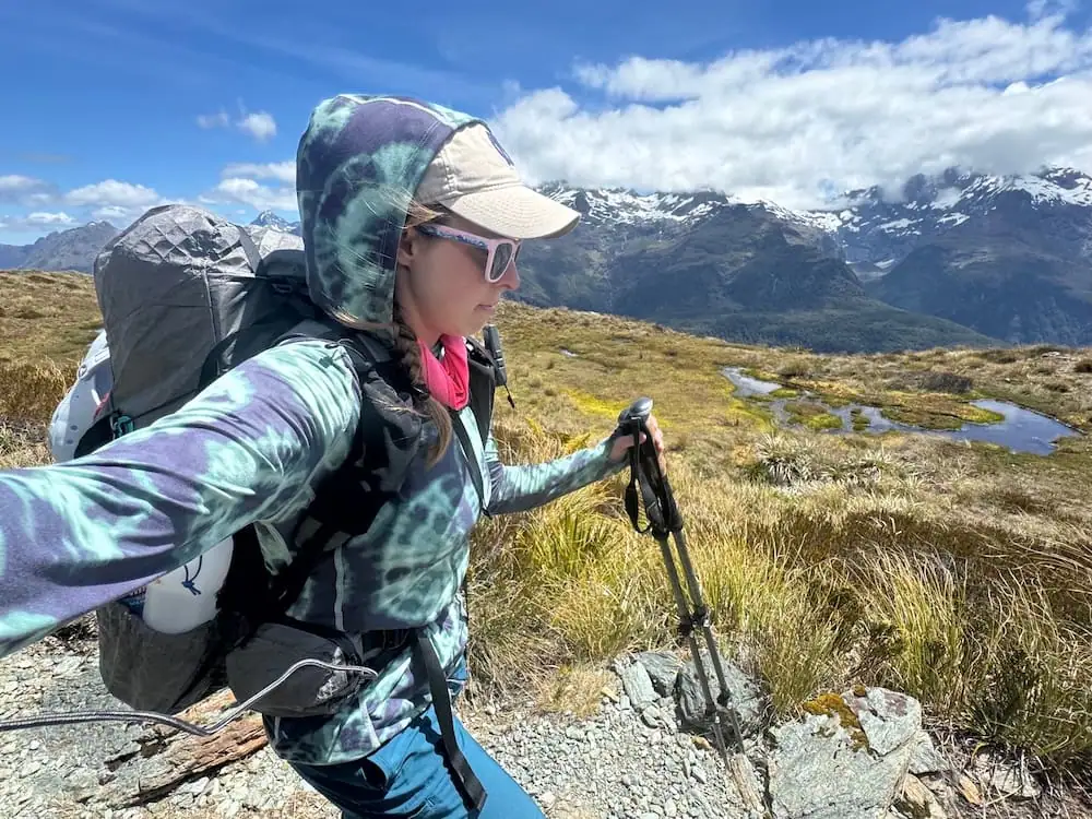Solo hiker carrying a backpack and trekking poles walks through open alpine terrain on the Routeburn Track. Snow capped mountains and winding streams stretch across the landscape beneath scattered clouds.