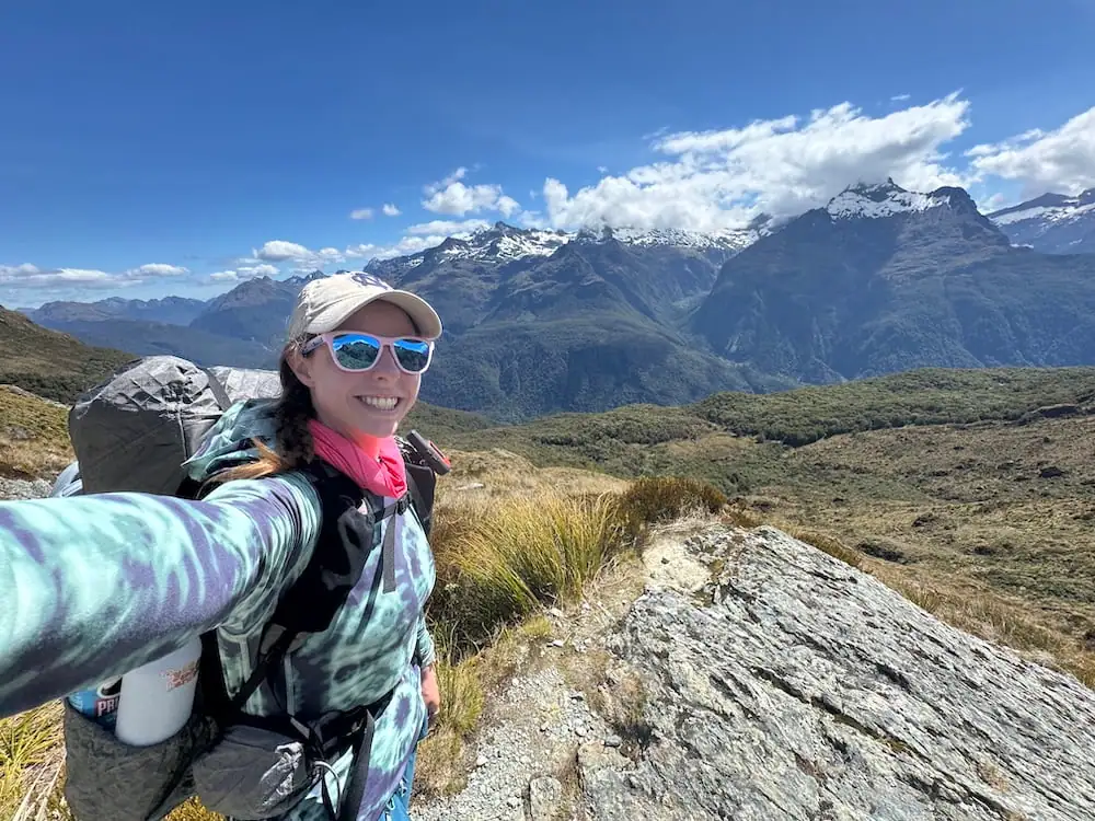 Smiling hiker with a backpack pauses for a selfie overlooking snow capped mountains and wide alpine valleys on the Routeburn Track. Bright sunshine highlights the open high country landscape.