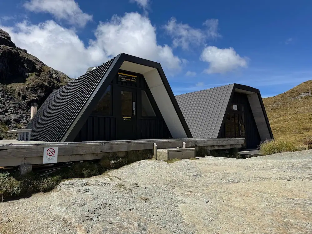 Dark A frame mountain emergency shelters surrounded by rocky alpine terrain and blue sky on the Routeburn Track. The modern shelter buildings sit in a remote high elevation setting.