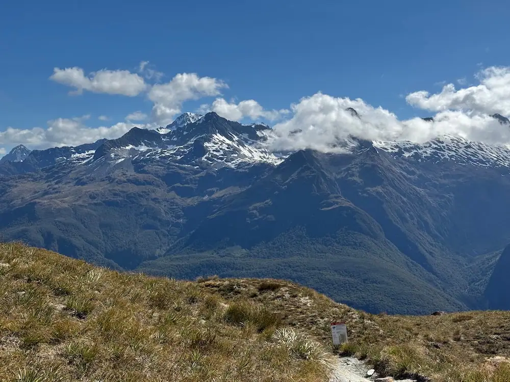 Snow capped mountain range rising above alpine grassland and a narrow trail on the Routeburn Track. Low clouds drift across the peaks under a bright blue sky, showing expansive views from higher elevations.