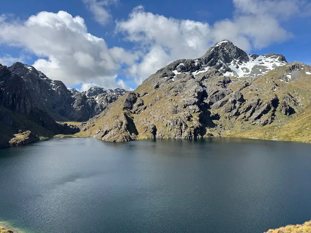Lake Harris surrounded by rocky mountains with patches of snow under a bright blue sky on the Routeburn Track. Calm water reflects the rugged peaks, showing the dramatic scenery hikers can expect along the trail.