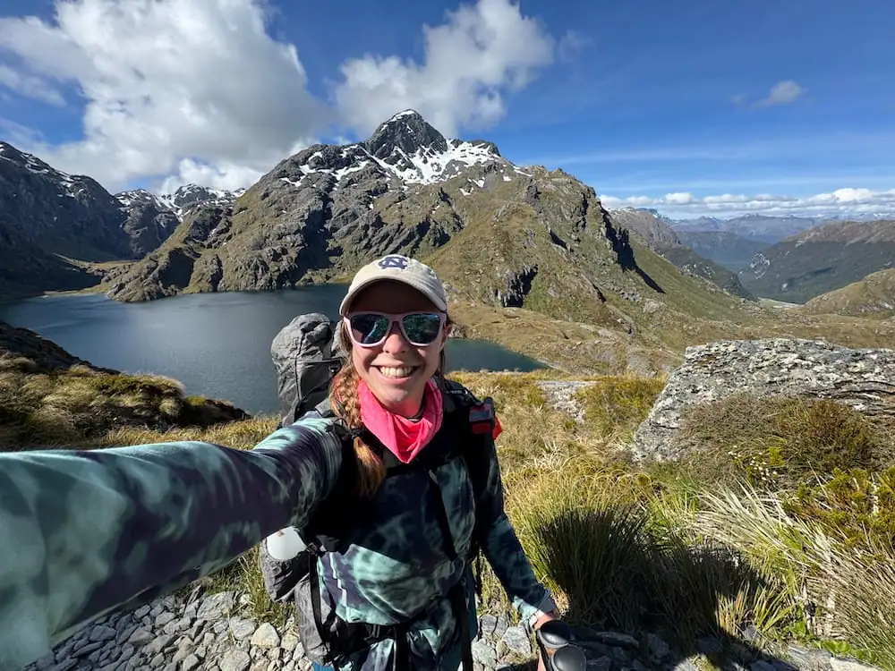 Smiling hiker with a backpack takes a selfie overlooking Lake Harris and snow capped mountain peaks on the Routeburn Track. Rocky cliffs and deep valleys stretch into the distance beneath scattered clouds.