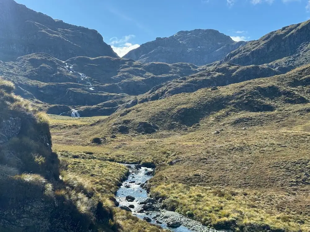 Small stream winding through open alpine grassland with rugged mountains rising in the background on the Routeburn Track. Thin waterfalls trickle down the rocky slopes under clear skies.