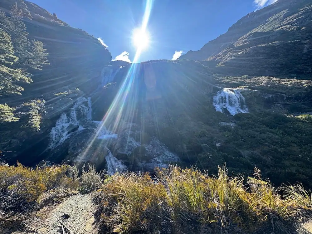 Sunlight beams across the cascading Routeburn Falls flowing down a rocky mountainside on the Routeburn Track. Golden alpine grasses line the foreground beneath a bright blue sky.