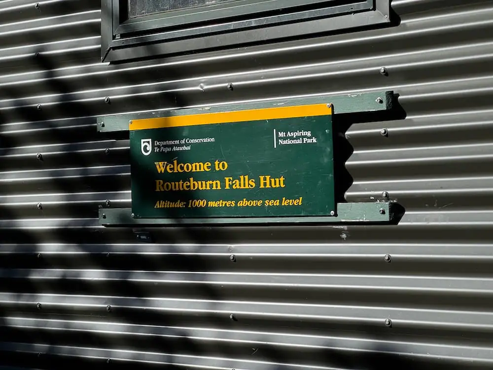 Green sign mounted on a corrugated metal hut wall reading "Welcome to Routeburn Falls Hut. Altitude 1000 metres above sea level." The hut sits along the Routeburn Track in Mt Aspiring National Park.