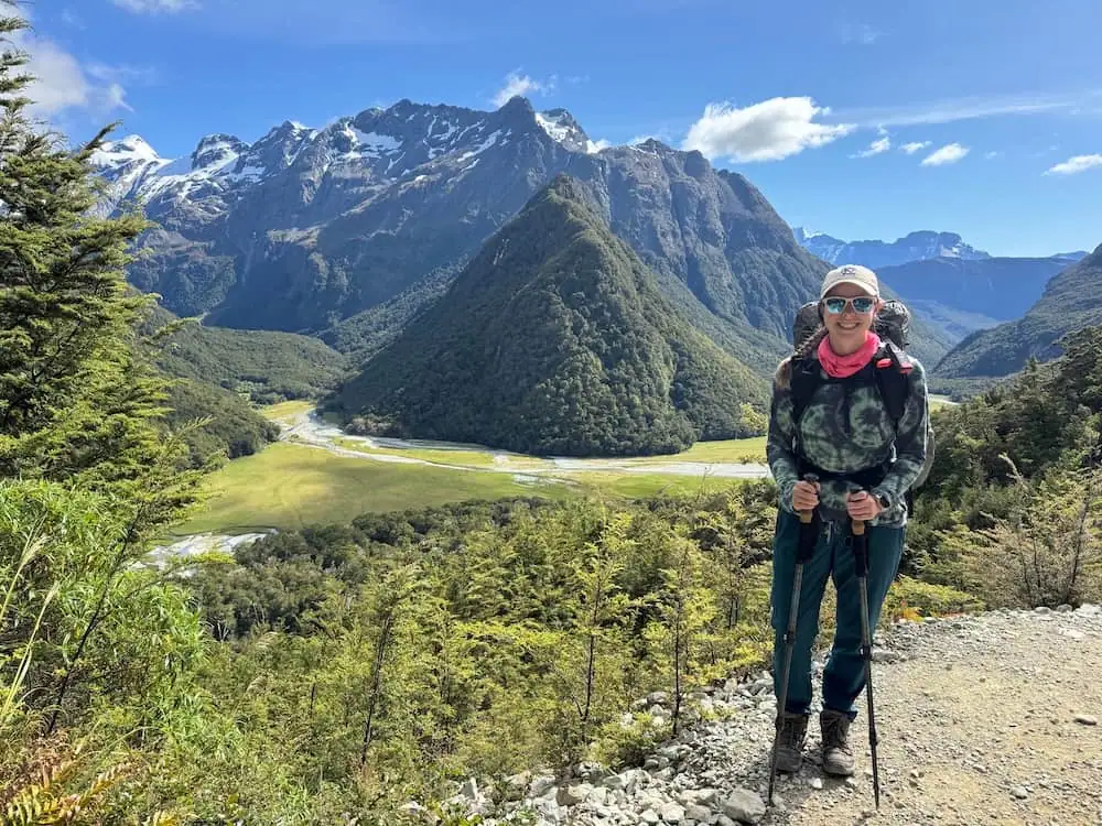 Hiker standing with trekking poles at a scenic overlook above a wide valley and rugged mountain peaks on the Routeburn Track. Bright sunshine and clear skies showcase expansive alpine views.