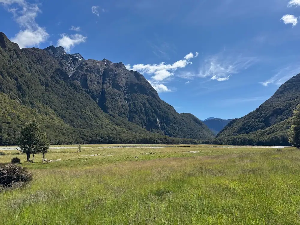 Wide grassy valley surrounded by steep forested mountains beneath a bright blue sky on the Routeburn Track. Sunlight stretches across the open landscape, showing the dramatic contrast between alpine peaks and flat valley floor.