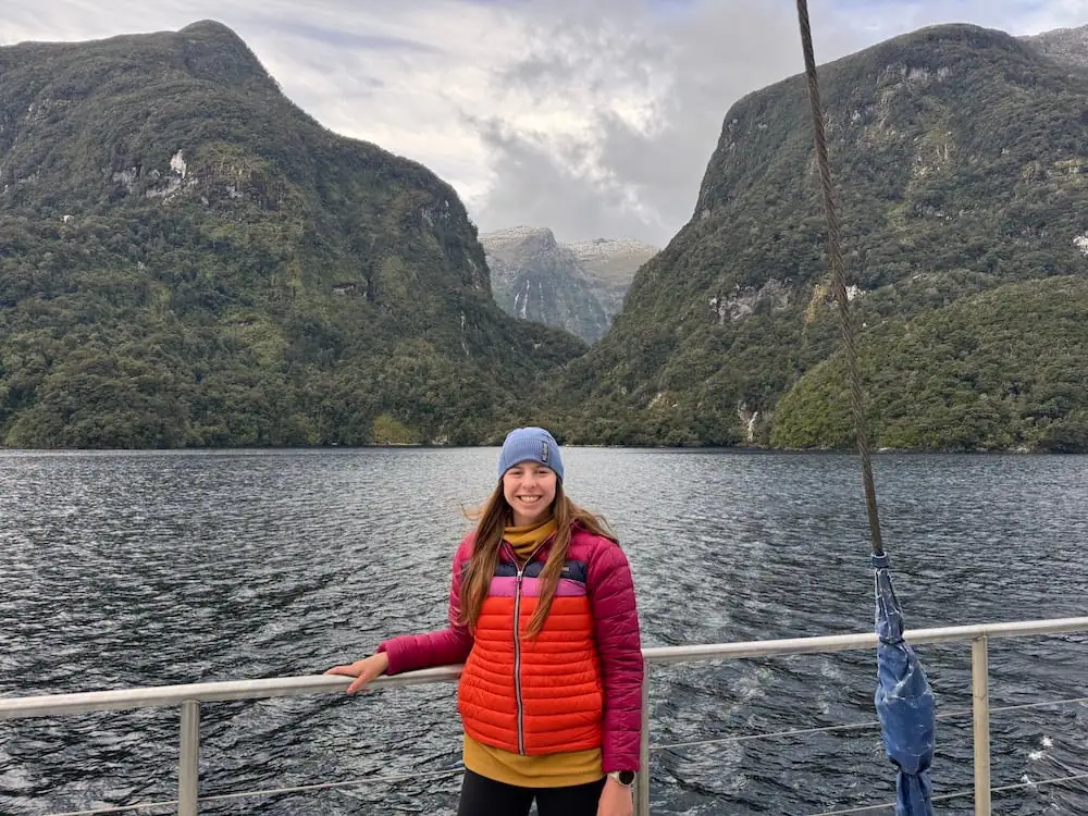 Smiling traveler in a bright jacket standing on a boat with towering forested cliffs and calm water behind her in Doubtful Sound. Misty mountain peaks rise between the valley walls beneath overcast skies.