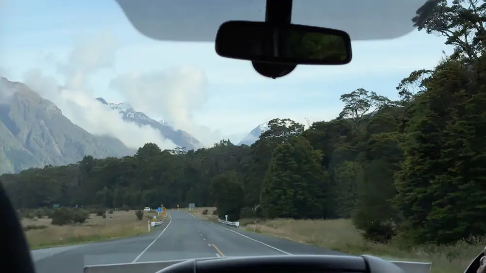 View through a vehicle windshield of a winding road lined with dense forest and misty mountains in the distance on the way to the Routeburn Track. Low clouds hang between steep peaks as the road curves into the valley.