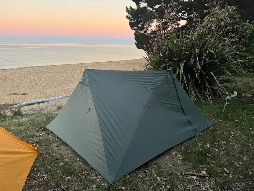 Lightweight hiking tent pitched near a quiet beach on the Abel Tasman Coast Track at sunset with soft pink skies over calm water. This campsite setup shows the type of overnight gear some hikers bring on Great Walks if they camp.