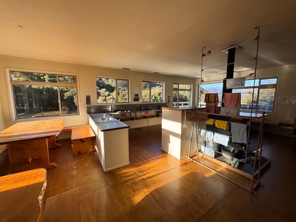 Bright and spacious hiking hut kitchen on the Heaphy Track with wooden tables, benches, drying racks, and large windows overlooking the outdoors. Shared hut facilities like this can help hikers decide what cooking and camp gear to include on an NZ Great Walks packing list.