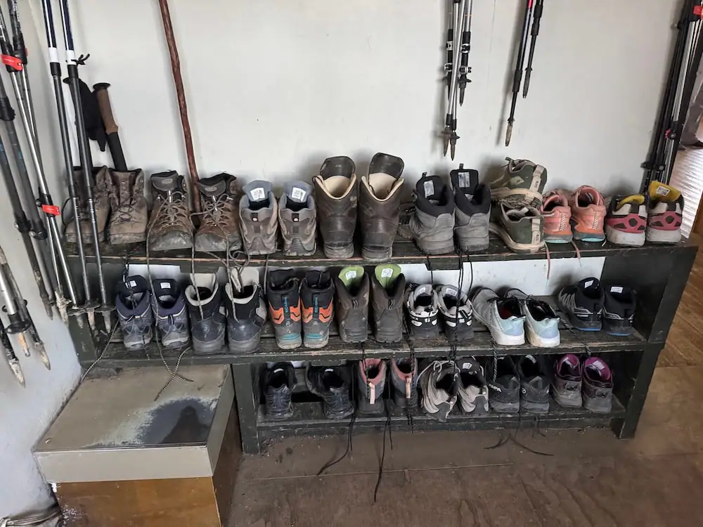 Muddy hiking boots, shoes, and trekking poles lined up on wooden shelves inside the Luxmore Hut on the Kepler Track. The variety of footwear and gear offers a realistic look at what people wear on the Great Walks in New Zealand.