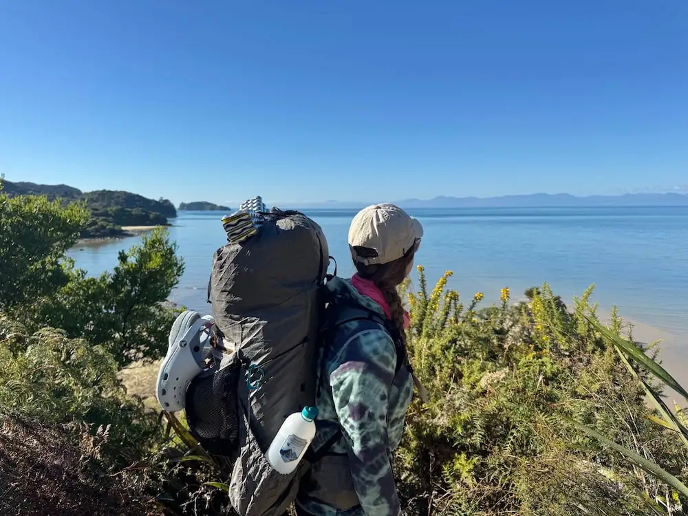 Mikala looking out over a calm coastline from the Abel Tasman Coast Track while carrying a large backpack filled with hiking gear on a sunny day. Visible items attached to her pack help show what hikers may carry on a Great Walk.