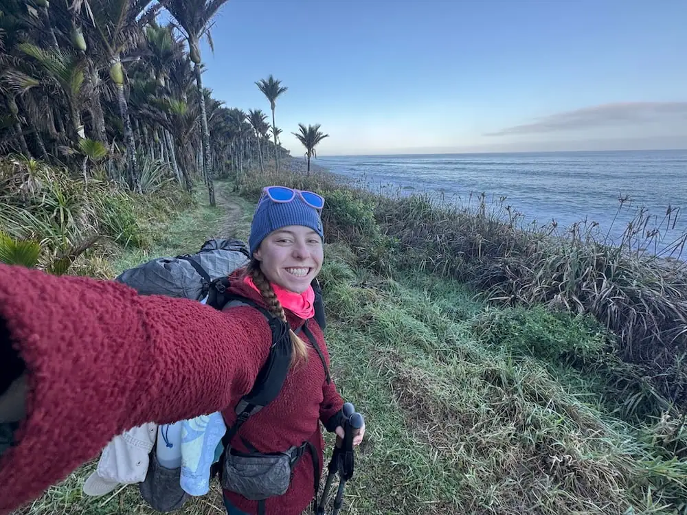 Mikala taking a selfie on the Heaphy Track beside the ocean shore lined with tall palm trees and native plants. Her cold weather layers and hiking setup demonstrate practical gear to consider packing for the Great Walks.