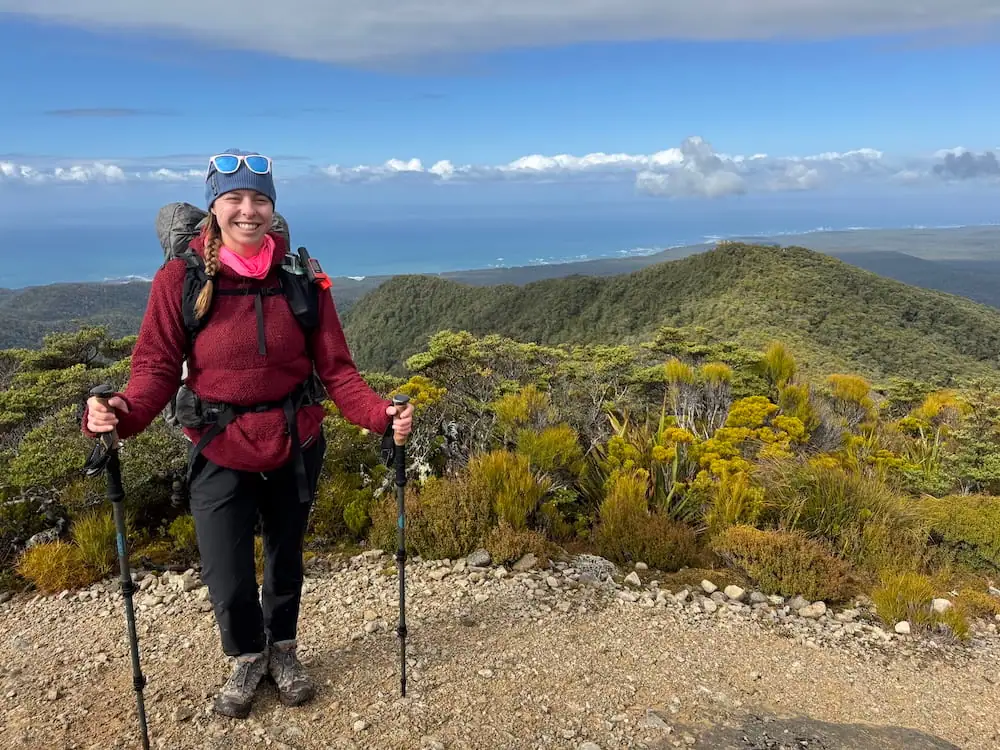Mikala smiling on a mountain summit on the Hump Ridge Track with trekking poles and a backpack while rolling green hills and ocean views stretch into the distance. Her layered clothing and hiking gear reflect practical choices for an NZ Great Walks packing list.