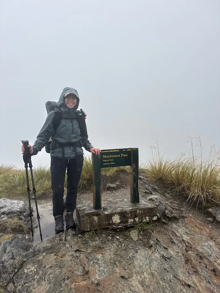 Mikala standing at the top of Mackinnon Pass on the Milford Track in rainy and foggy weather while wearing a waterproof jacket and carrying trekking poles. Wet weather gear like rain layers is an important part of an NZ Great Walks packing list.