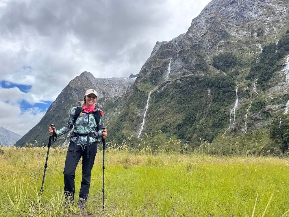 Mikala standing in a grassy valley on the Milford Track surrounded by steep mountains and tall waterfalls while wearing hiking gear and carrying trekking poles. This landscape highlights the kind of alpine scenery hikers should anticipate packing for on the Great Walks.