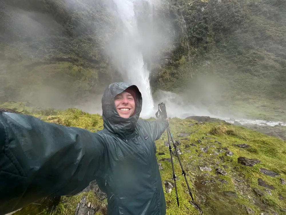 Mikala taking a selfie in heavy rain beside Sutherland Falls while wearing full waterproof rain gear and holding trekking poles. Wet conditions like this highlight why rain protection is essential on an NZ Great Walks packing list.