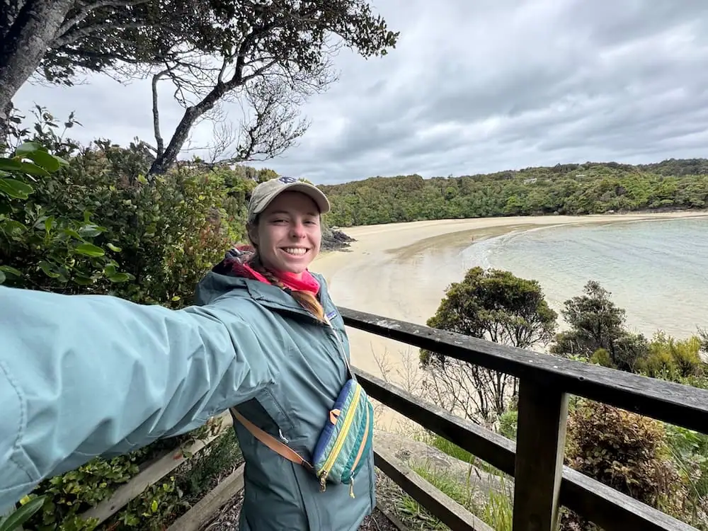 Mikala smiles at the camera from a wooden lookout above a Bathing Beach, wearing a rain jacket and cap beneath a moody overcast sky. Staying at an accommodation in Oban is just a short walk from here.