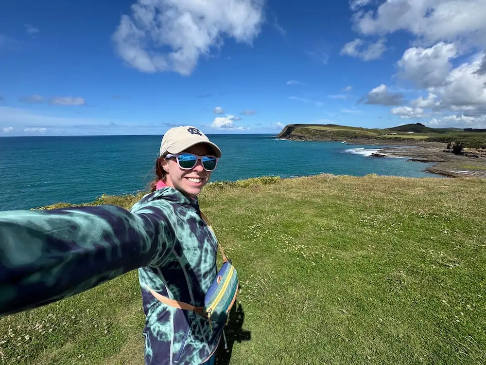 Mikala smiles for a selfie at Curio Bay Cliffs in the Catlins, wearing sunglasses and a baseball cap with turquoise ocean, rugged coastline, and rolling green headlands behind her. This scenic stop was part of the drive to Invercargill Airport before continuing the journey to Stewart Island.