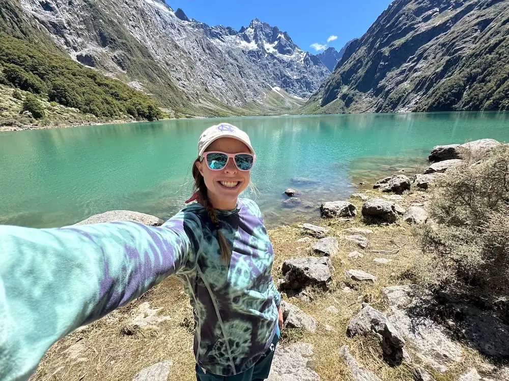 Mikala smiles for a selfie at the edge of the turquoise waters of Lake Marian, surrounded by steep rocky peaks and alpine scenery at the end of the Lake Marian Track. Hiking to this stunning lake is one of the most rewarding things to do in the Te Anau area for outdoor lovers.