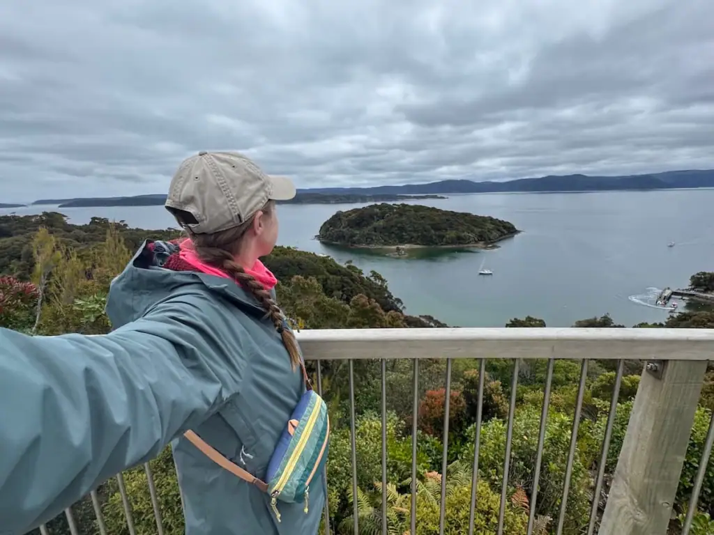 Mikala stands at a scenic lookout at Observation Rock in a rain jacket and baseball cap, gazing over Stewart Island's sheltered bays and forested islets beneath a dramatic cloudy sky.