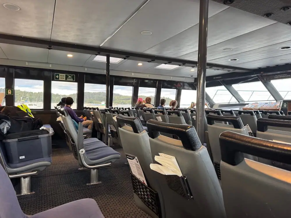 Interior of the passenger cabin on the boat used for the Stewart Island kiwi tour, with rows of grey seats facing large windows overlooking the water. A handful of passengers sit quietly as the vessel travels toward the evening wildlife experience.
