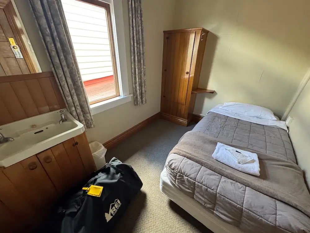 Compact single guest room at the South Sea Hotel on Stewart Island with a neatly made bed, wooden wardrobe, bedside shelf, and in-room sink beneath a sunlit window. A folded towel rests on the bed, highlighting the practical amenities available for solo travelers and hikers.