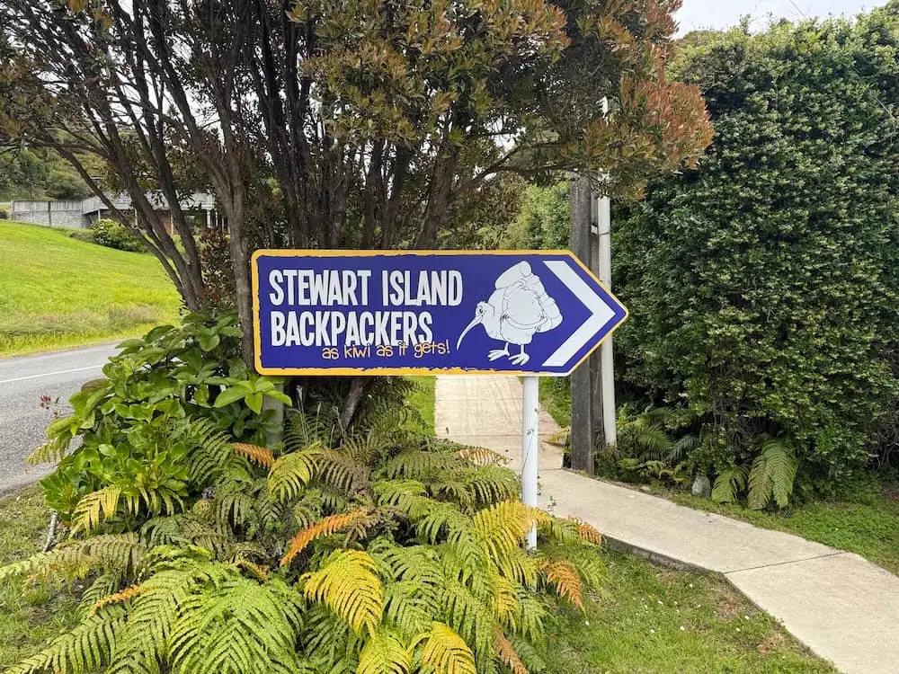 Blue roadside sign reading "STEWART ISLAND BACKPACKERS as kiwi as it gets!" with an illustration of a kiwi bird and a directional arrow pointing toward the property. Surrounded by native plants and greenery, the sign marks the location of this Stewart Island accommodation for arriving guests.