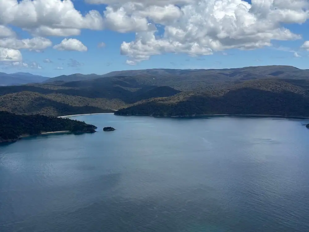 Aerial view of Stewart Island showing calm blue bays bordered by densely forested hills beneath large white clouds. Seeing the island emerge below the plane offers a glimpse of the final approach for travelers flying to Stewart Island from the South Island.