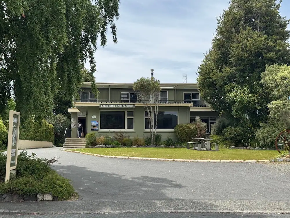 Front entrance of Te Anau Lakefront Backpackers with trees and landscaped grounds surrounding the building. The sign above the entrance reads "Lakefront Backpackers."