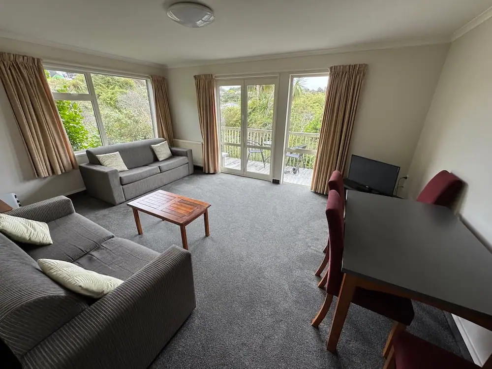 Spacious living area at Stewart Island accommodation The Bay Motel featuring two grey sofas, a wooden coffee table, a dining table with red chairs, and large windows and patio doors overlooking lush native greenery. The bright communal space offers guests a comfortable place to relax while enjoying views of Stewart Island's surrounding bush.