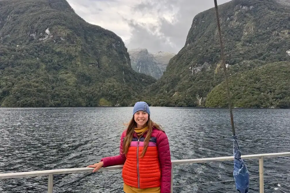 Smiling traveler in a bright jacket standing on a boat with towering forested cliffs and calm water behind her in Doubtful Sound. Misty mountain peaks rise between the valley walls beneath overcast skies.
