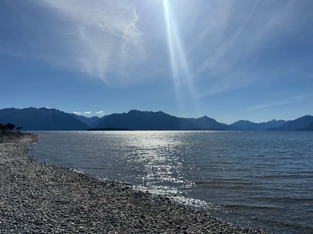 Sunlight sparkles across the lake at Henry Creek Campsite with a rocky shoreline in the foreground and mountain ranges stretching across the horizon under a clear blue sky. The peaceful lakeside view shows the natural scenery that makes camping near Te Anau a memorable accommodation experience.