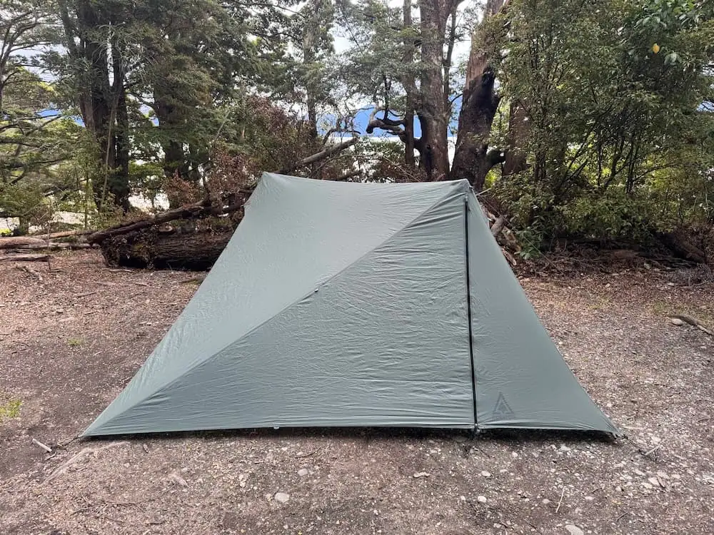 Lightweight backpacking tent pitched on a gravel campsite at Henry Creek Campsite, surrounded by mature native trees with glimpses of the lake beyond. This simple campsite is a scenic Te Anau accommodation choice for campers exploring Fiordland.