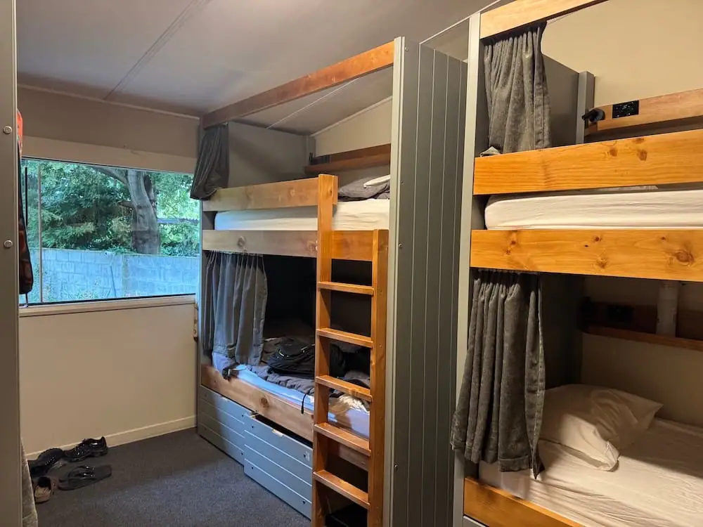 Shared hostel bunk room at Te Anau Lakefront Backpackers with sturdy wooden bunk beds, privacy curtains, under bed storage, and a large window overlooking trees. This comfortable dorm style setup is a budget friendly Te Anau accommodation option for travelers exploring Fiordland.