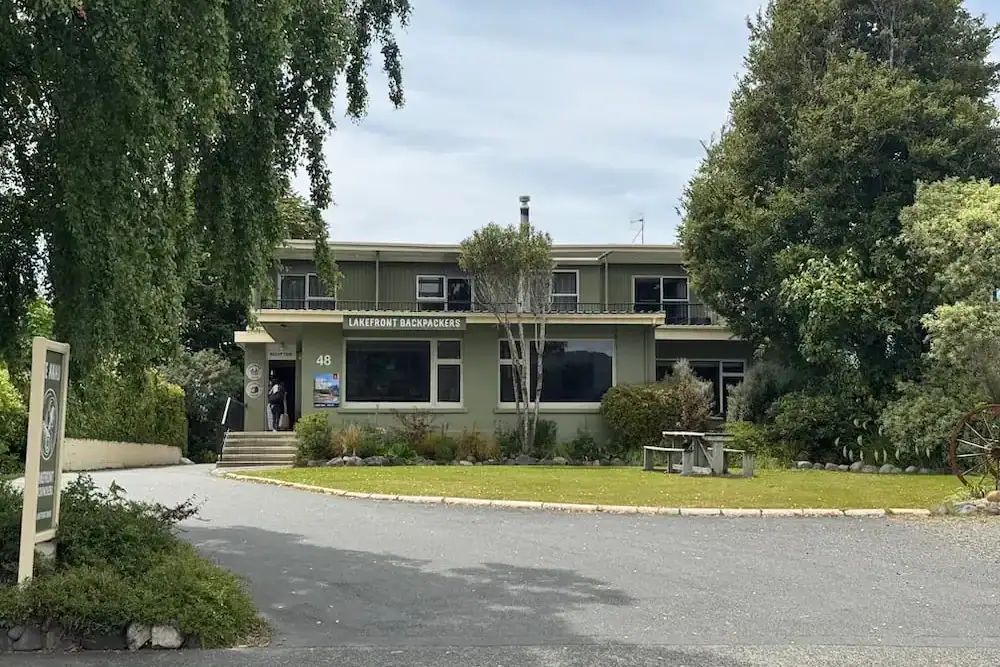 Front entrance of Te Anau Lakefront Backpackers with trees and landscaped grounds surrounding the building. The sign above the entrance reads "Lakefront Backpackers."