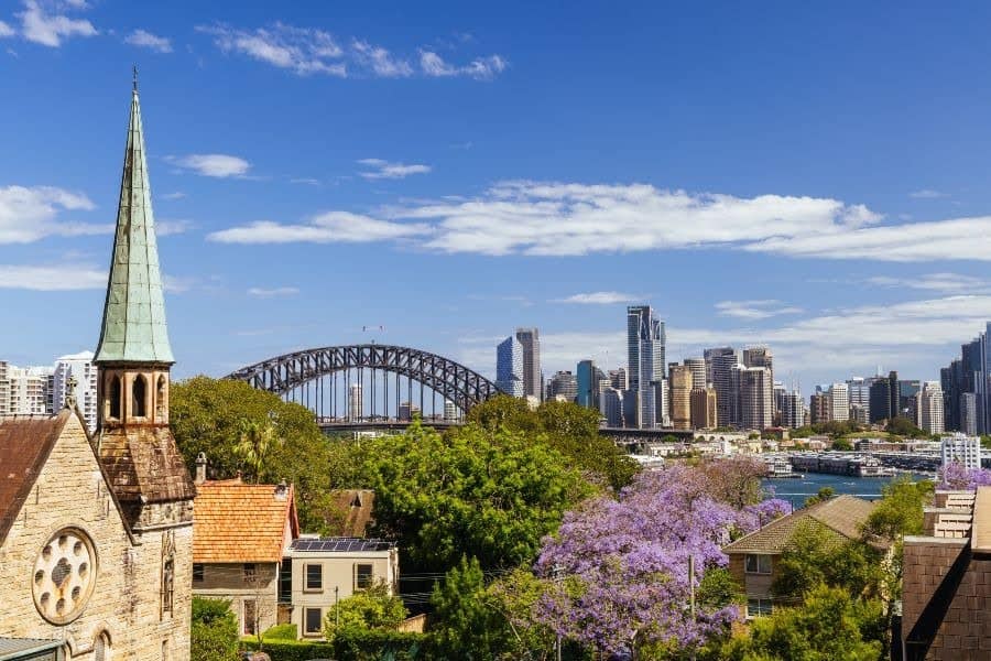 Sydney Harbour Bridge and skyline, with a church spire and jacaranda trees.