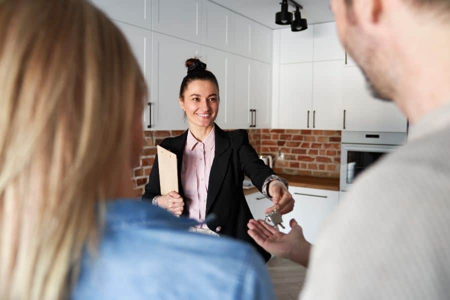 Smiling real estate agent handing house keys to tenants inside a modern kitchen.