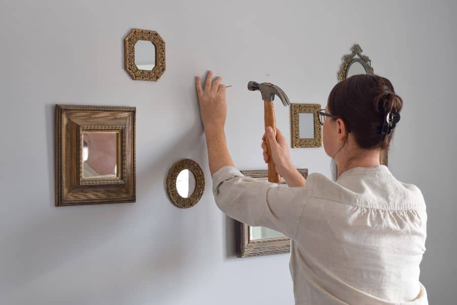 Woman using a hammer to drive a nail into a wall while arranging framed mirrors in her home. 