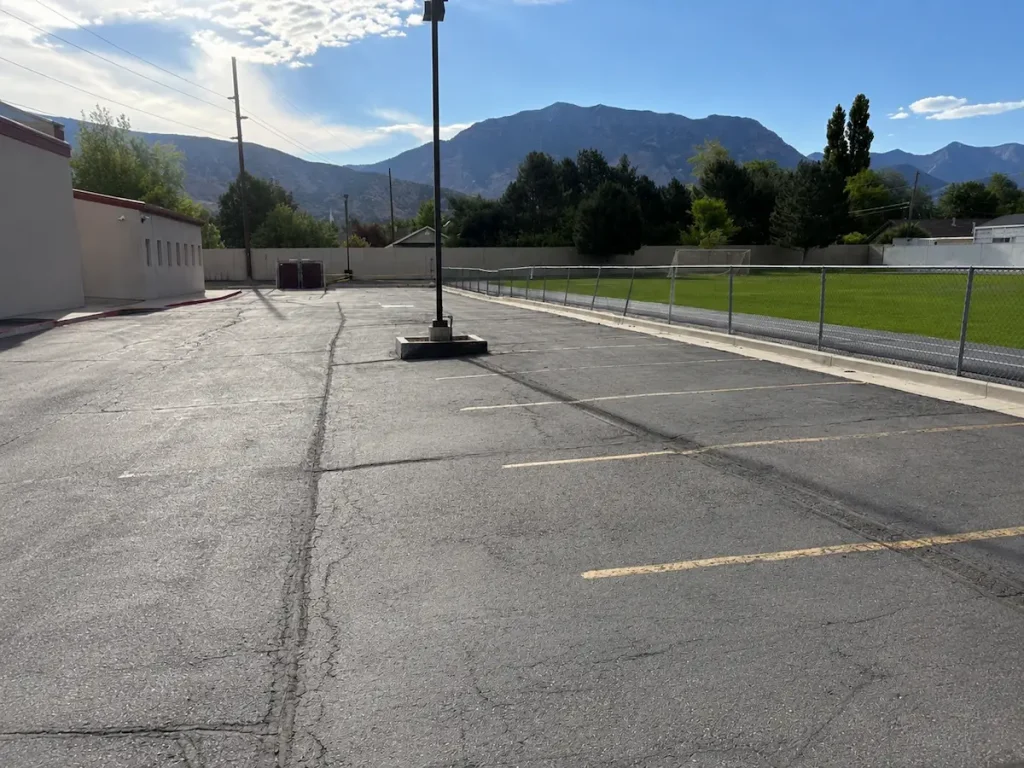 Before photo of faded parking lot with worn asphalt, visible cracks, and faded parking stall striping at Timpanogos Academy in Lindon, Utah before sealcoating.