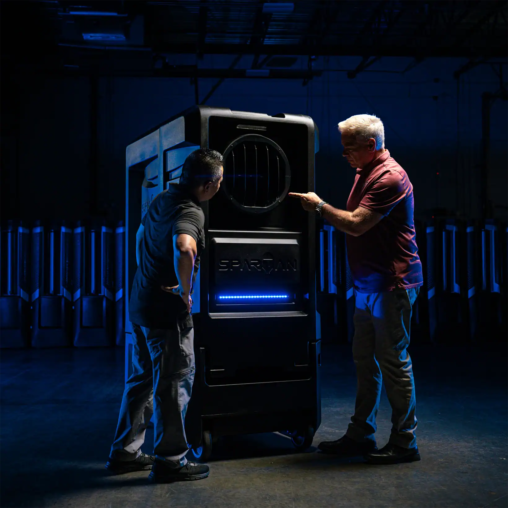 Two Vector team members inspecting Spartan evaporative cooler in a dimly lit industrial warehouse, highlighting its large vent and illuminated control panel.